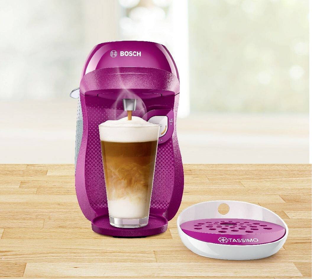 'Magenta-farbene Tassimo Kaffeemaschine mit Milchschaum-Latte im Vordergrund auf einem Holztisch.'