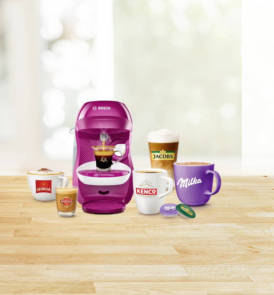 Eine rosafarbene Bosch Tassimo Kaffeemaschine mit einer gefüllten Tasse in der Mitte. Verschiedene Tassen und Marken im Vordergrund.