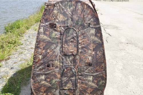 Ein Zelt in Tarnfarbe steht auf einer gepflasterten Fläche neben einem Fluss. Das Zelt hat zwei Fenster und ist für Outdoor-Aktivitäten geeignet.