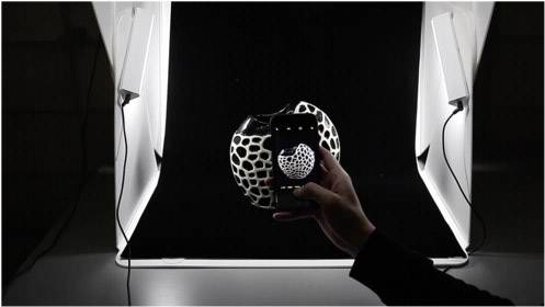 Eine Hand hält ein Smartphone, das ein Foto von einem kugelförmigen Objekt mit schwarzem und weißem Muster unter Beleuchtung aufnimmt.