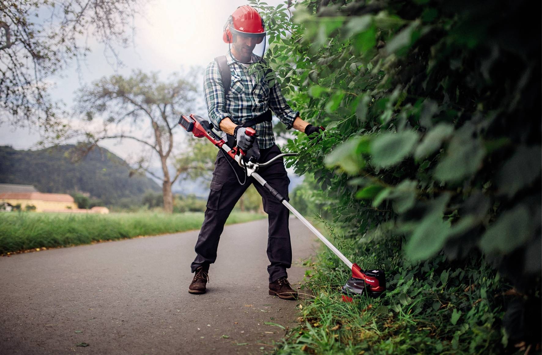 Ein Mann mit Schutzhelm und Visier schneidet mit einem Rasentrimmer Gras am Wegrand entlang eines Weges in einer ländlichen Umgebung.