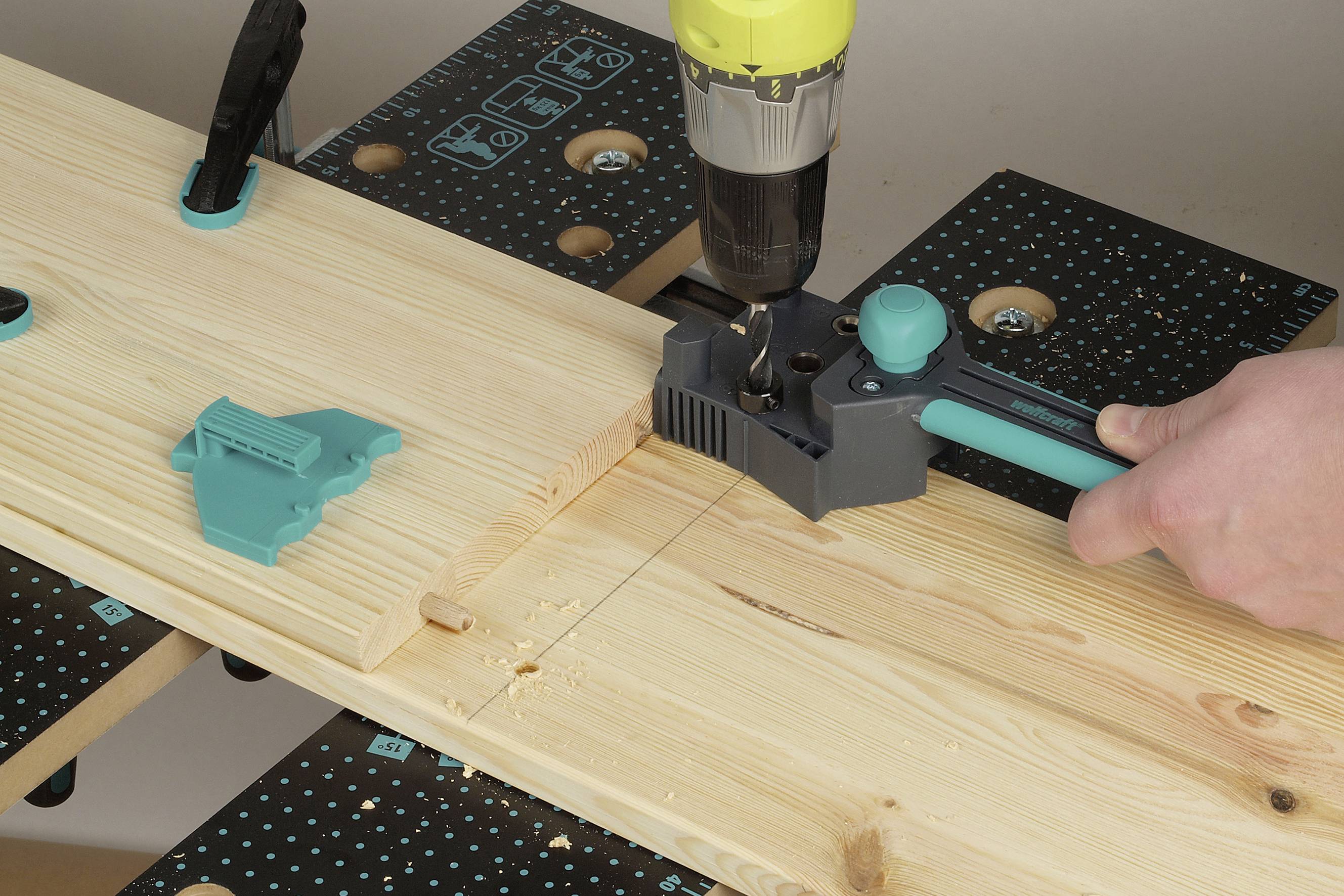 Eine Person bohrt ein Loch in ein Holzbrett, das in einem Spannwerkzeug gesichert ist. Markierungen auf dem Holz zeigen das Bohrmuster.