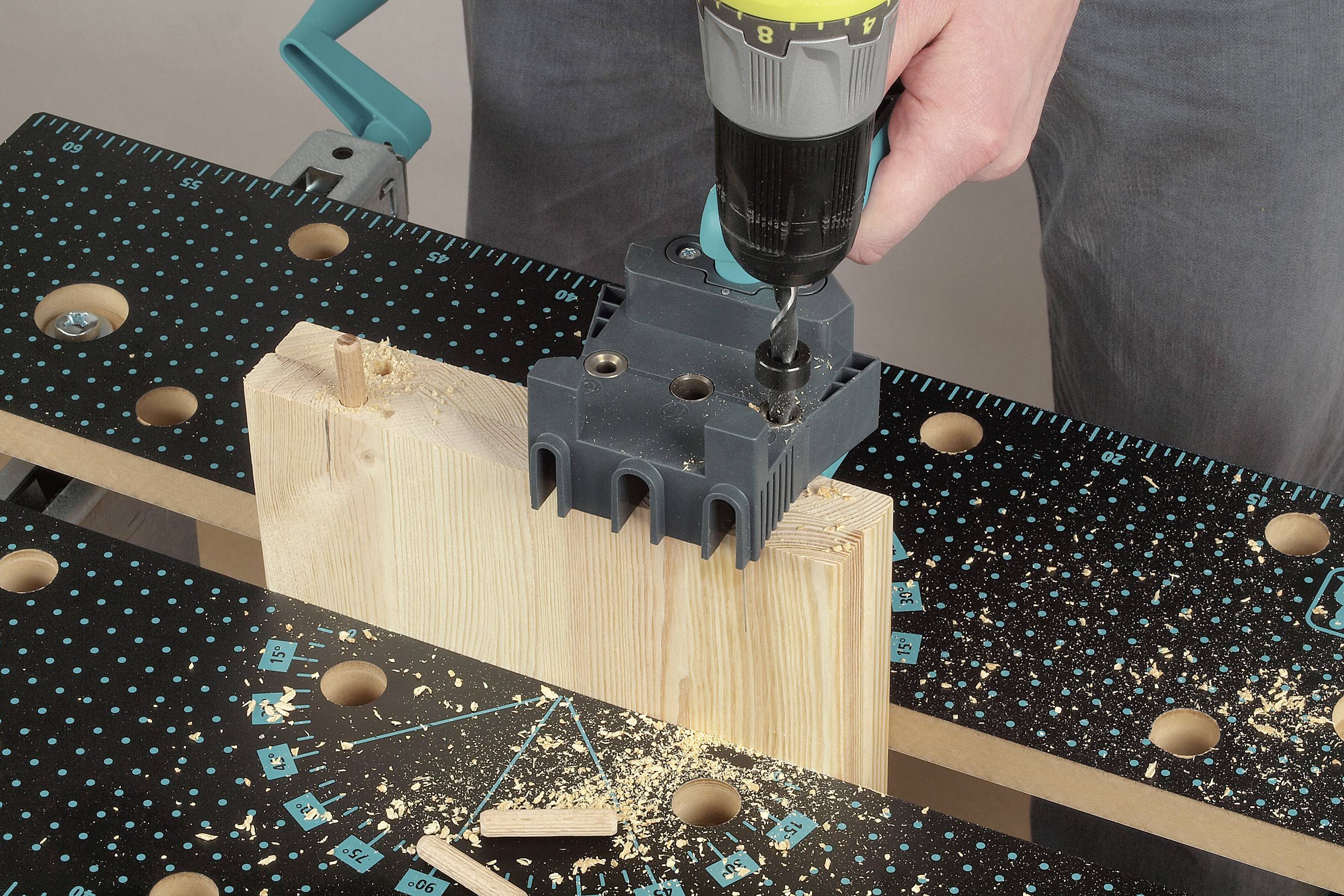 Eine Person bohrt mit einer Bohrmaschine in ein Holzstück, das in einer Werkbank eingespannt ist. Holzspäne sind auf der Werkbank sichtbar.