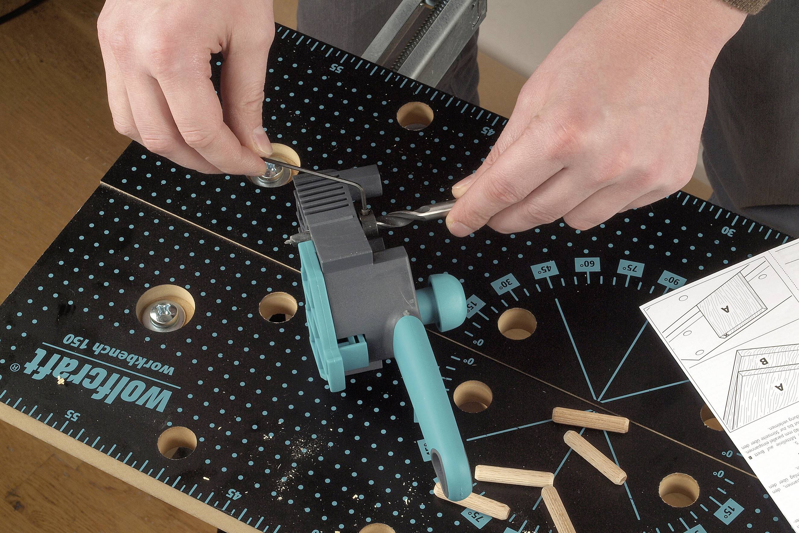 'Person montiert eine Fräsvorrichtung auf einem Arbeitstisch. Holzstücke und ein Bauplan liegen daneben.'