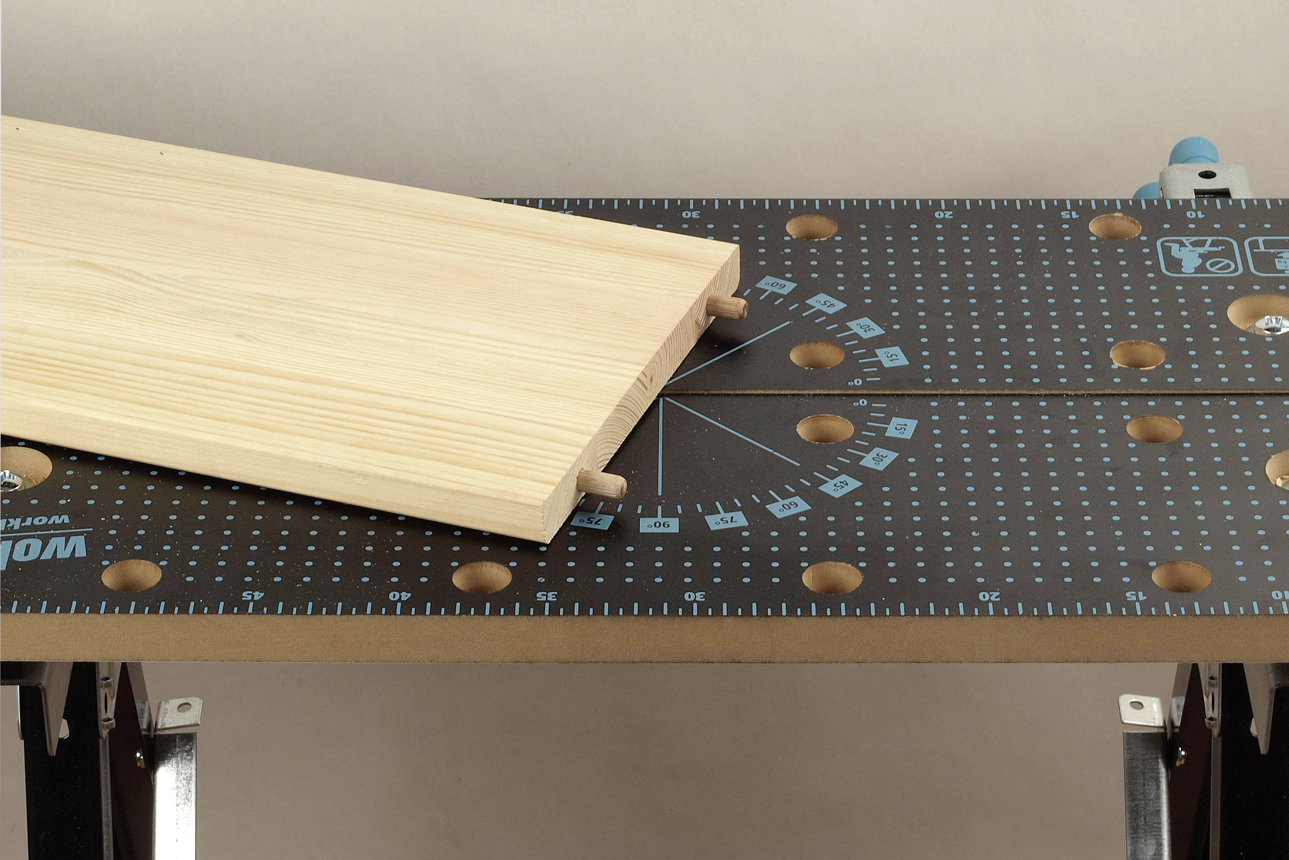 Holzbrett mit Dübeln liegt auf einem Werktisch mit Maßlinien und Bohrlöchern, bereit zur Montage oder Bearbeitung.