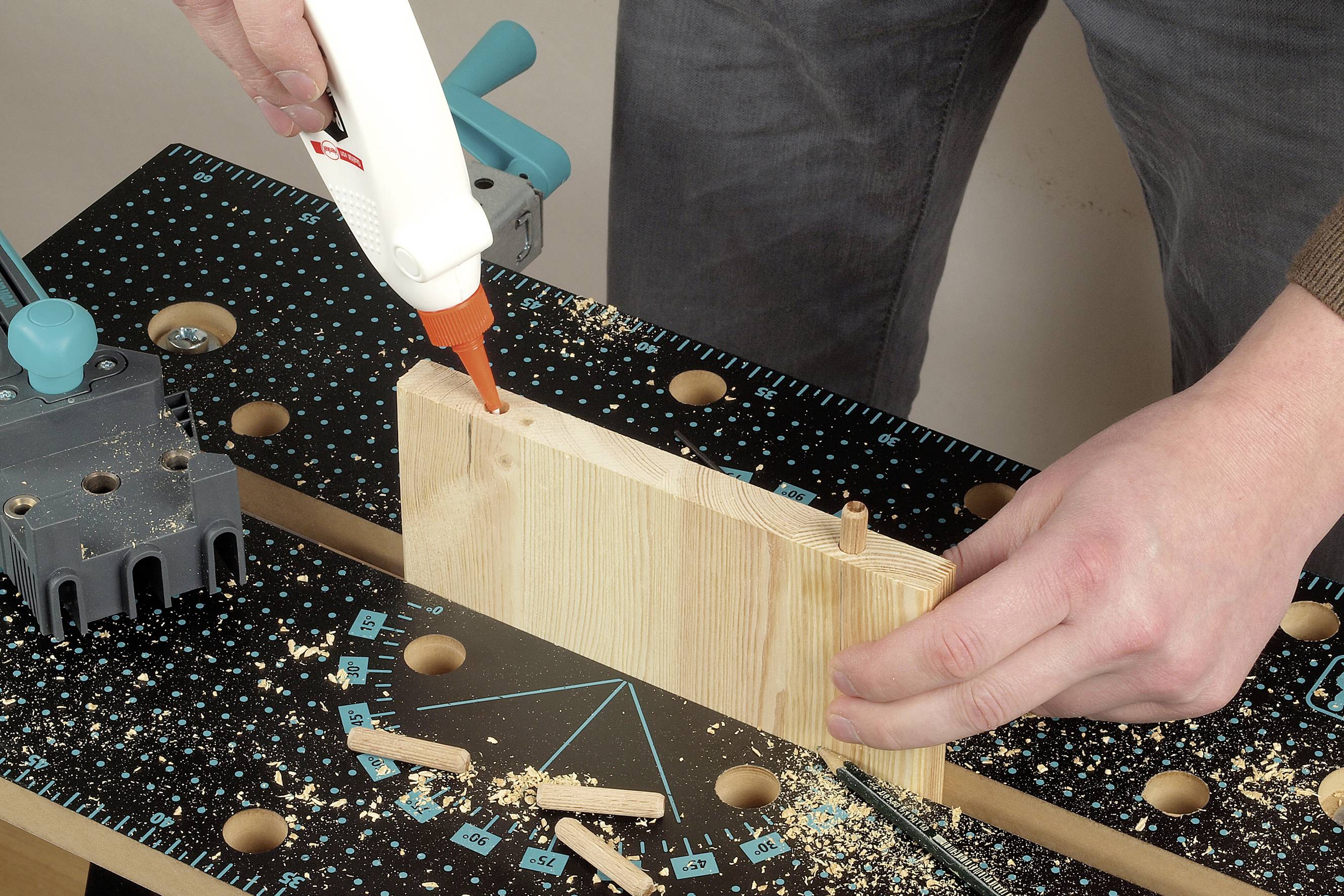 Eine Person trägt Holzleim auf das Ende eines Holzbretts auf, das in einer Werkbank festgeklemmt ist. Umgebende Holzspäne sind sichtbar.