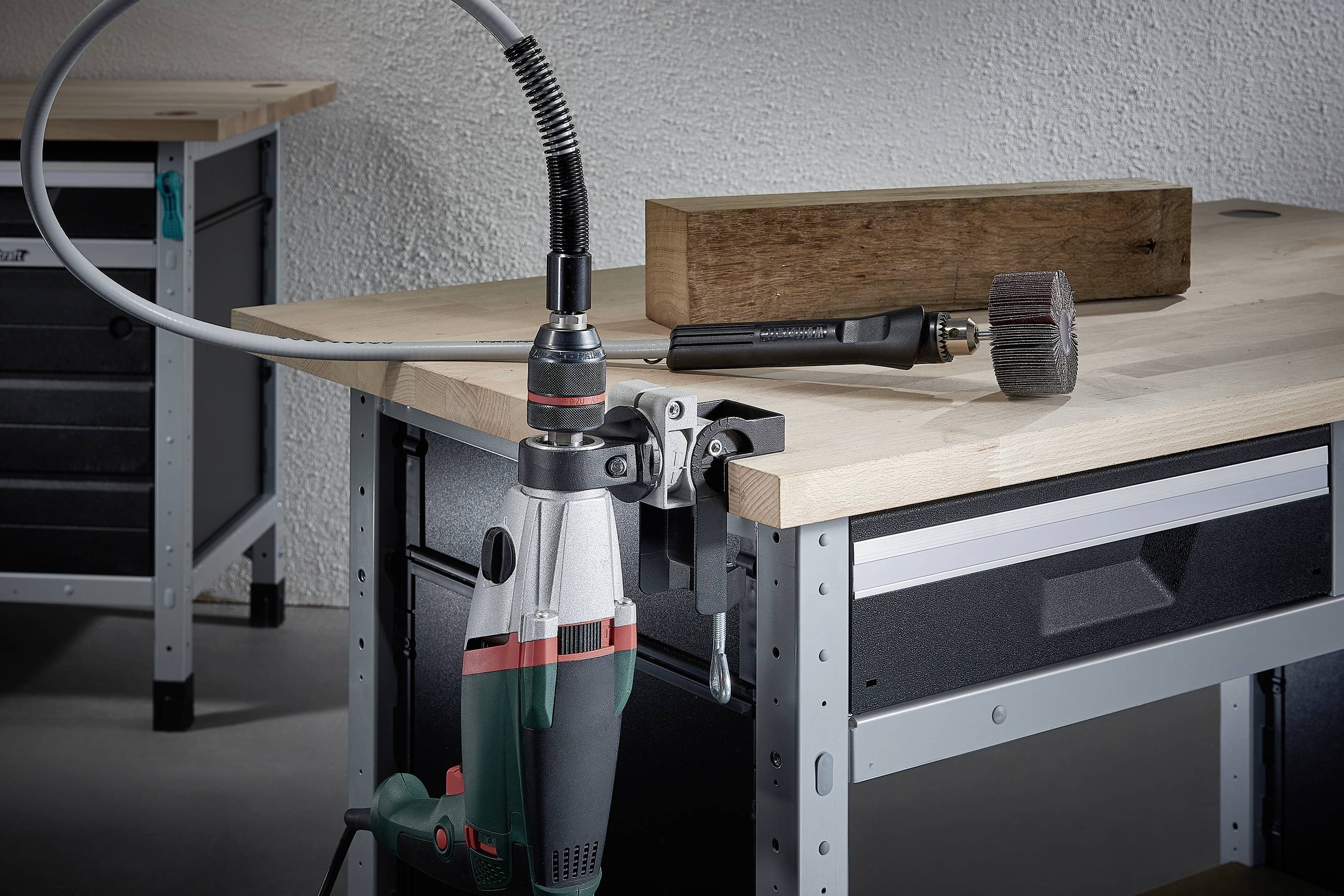 In einer Werkstatt steht eine Bohrmaschine auf einem Tisch, daneben ein Holzblock und ein großes Schraubenwerkzeug.