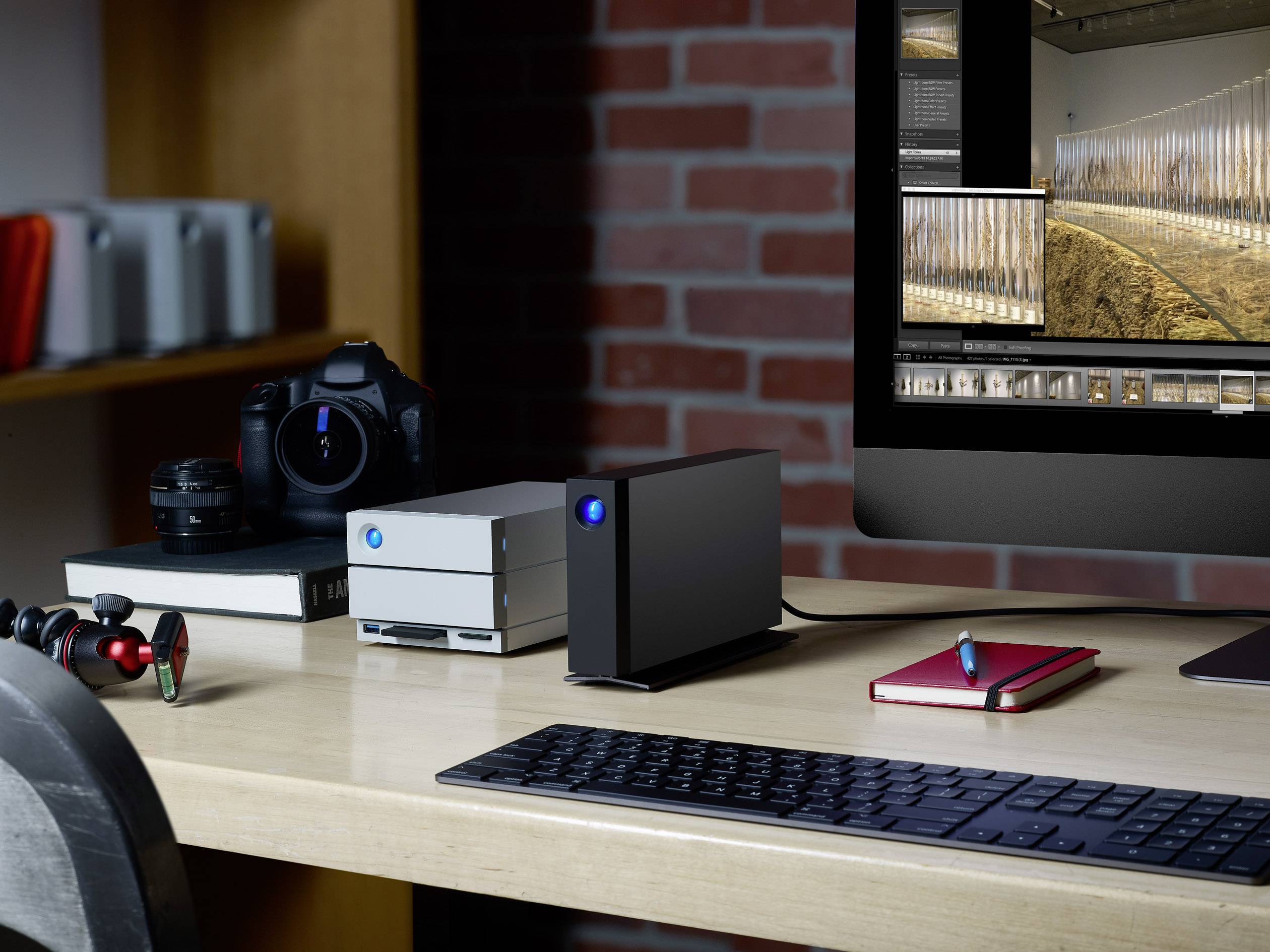 Ein Schreibtisch mit einem Monitor, auf dem Bildbearbeitungssoftware geöffnet ist, umgeben von Festplatten, einer Kamera und einem Notizbuch.