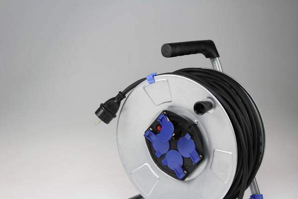 Ein silbernes Kabeltrommel mit schwarzem Griff und Kabel. Vier blaue Steckdosen in der Mitte. Die Kabeltrommel steht auf einem grauen Hintergrund.