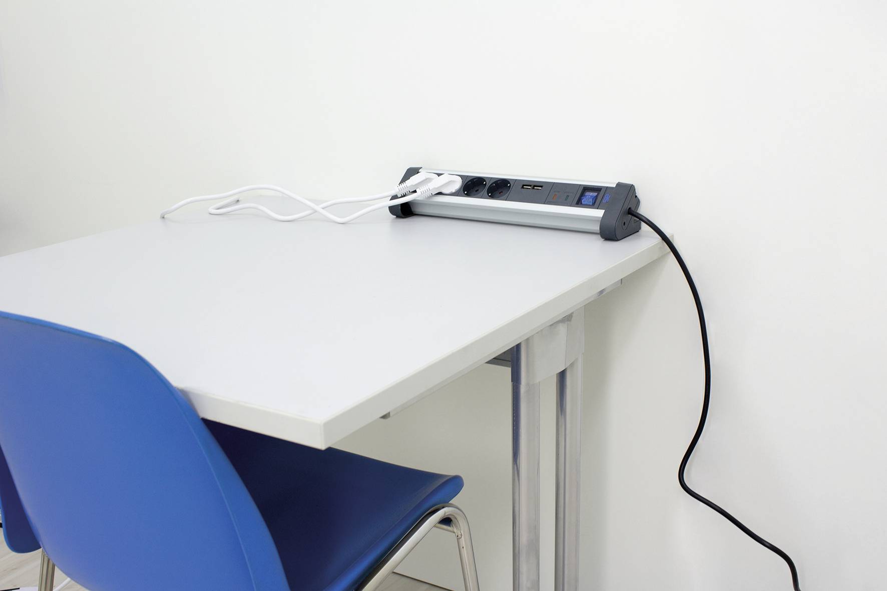 Ein blauer Stuhl steht neben einem weißen Tisch, auf dem eine Steckdosenleiste mit eingesteckten weißen Kabeln liegt.
