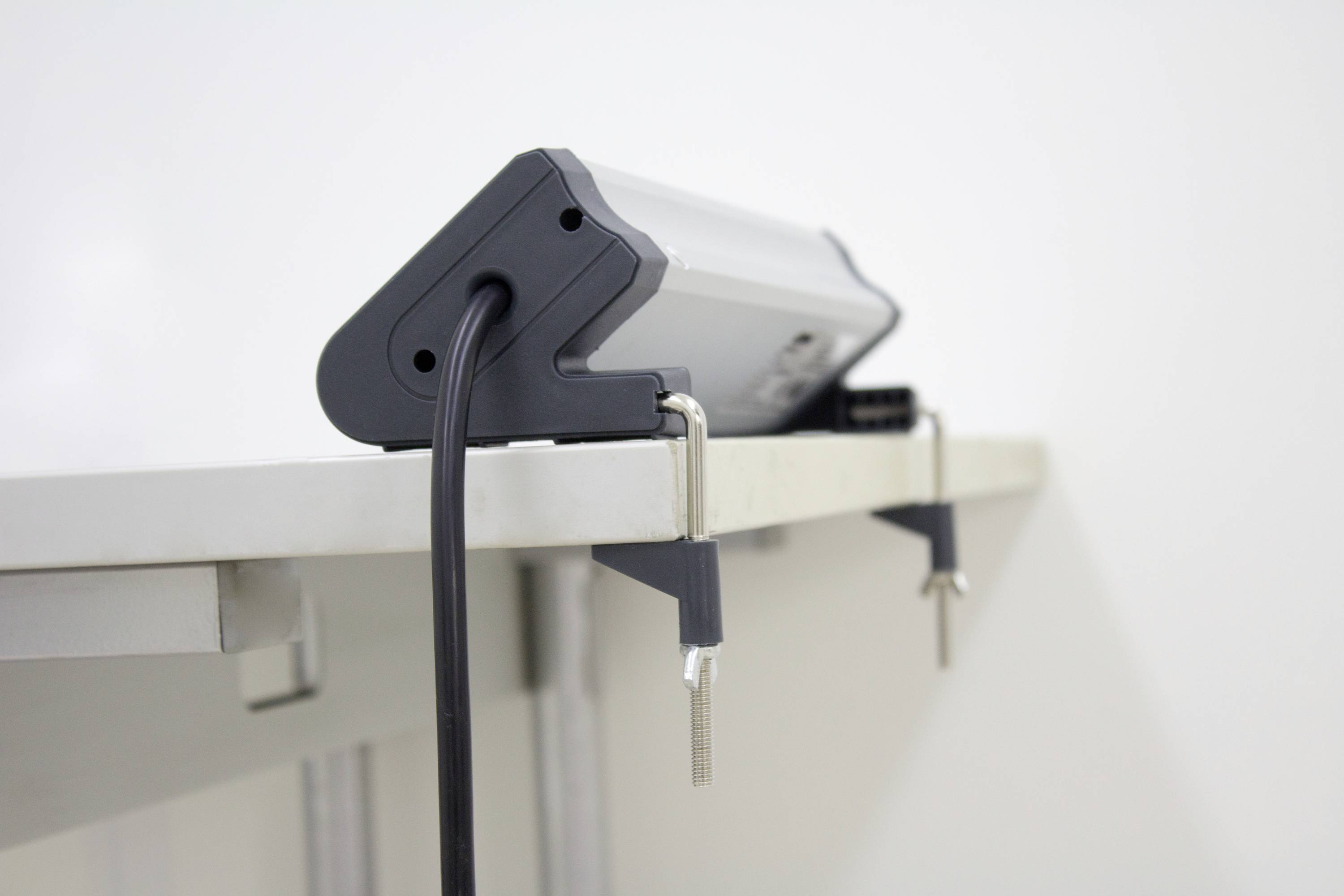 Leuchte mit schwarzem Kabel, befestigt an der Kante eines weißen Tisches mit Metallklemmen.