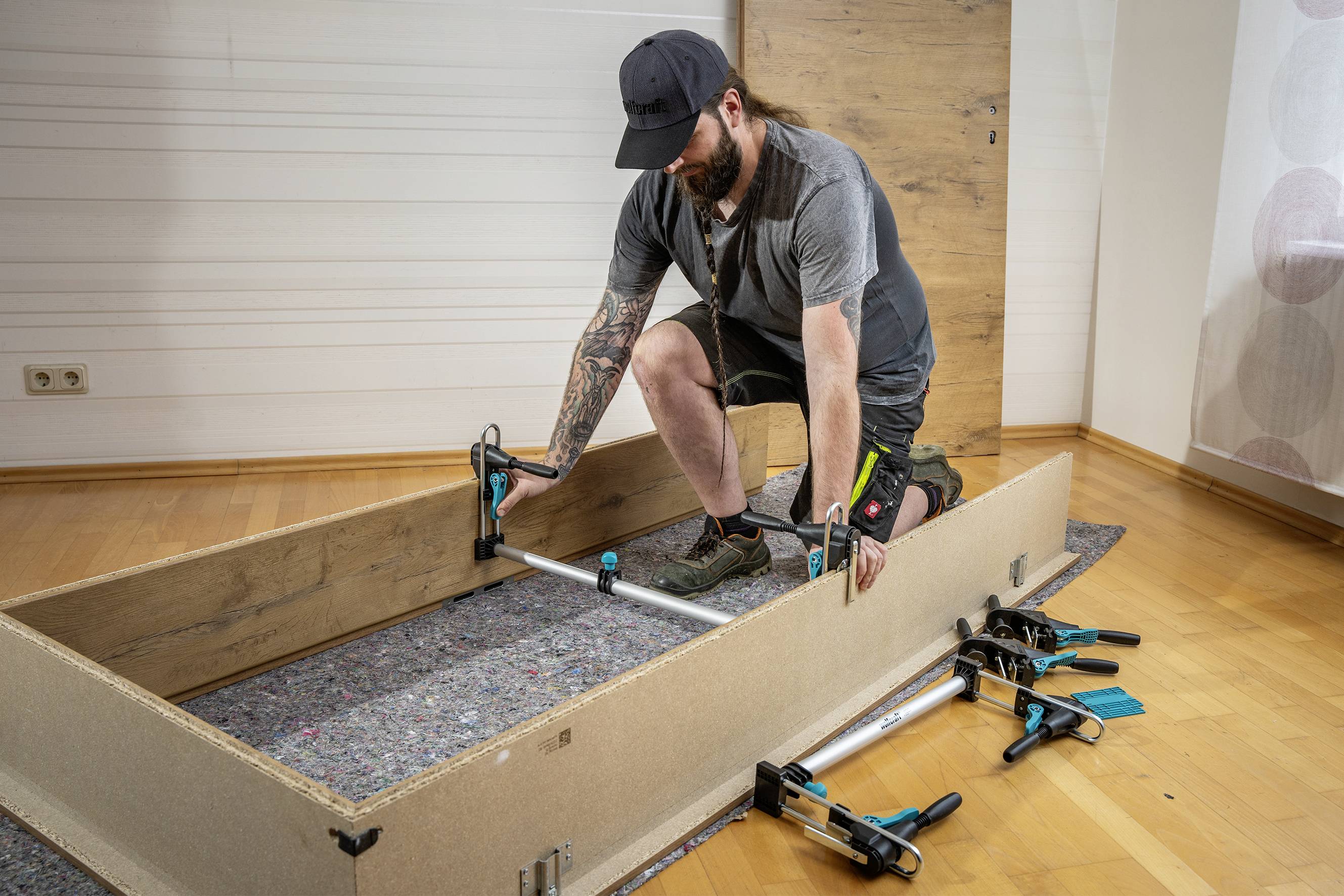 Mann in Handwerkerkleidung montiert ein Möbelstück mit Klemmen. Kniebeugend arbeitet er an einem rechteckigen Holzrahmen auf einem Teppich.