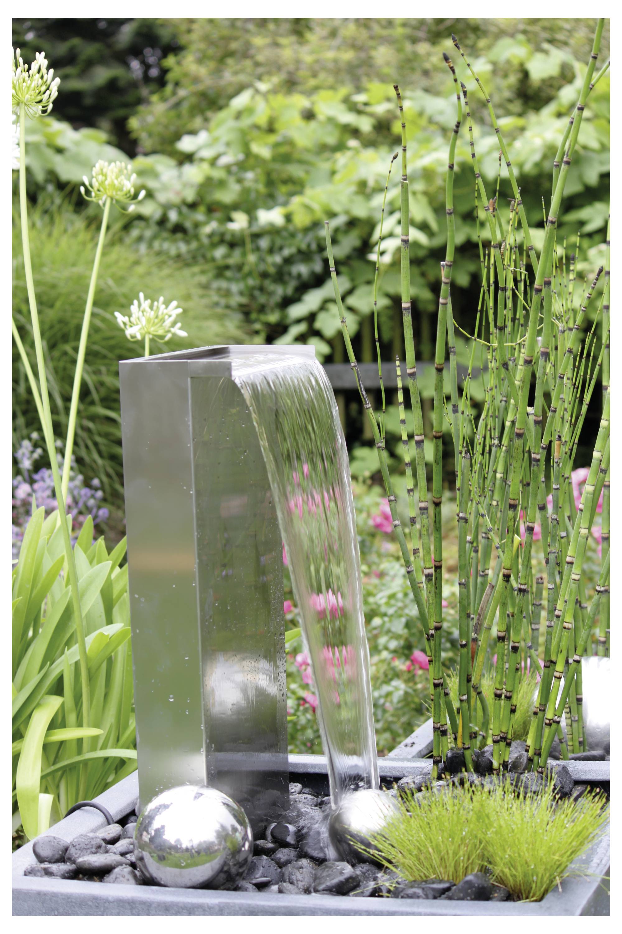 Ein moderner Gartenbrunnen mit Wasser, das aus einer silbernen Metallmündung fließt, umgeben von grünen Pflanzen und Blumen, der die Außendekoration aufwertet.