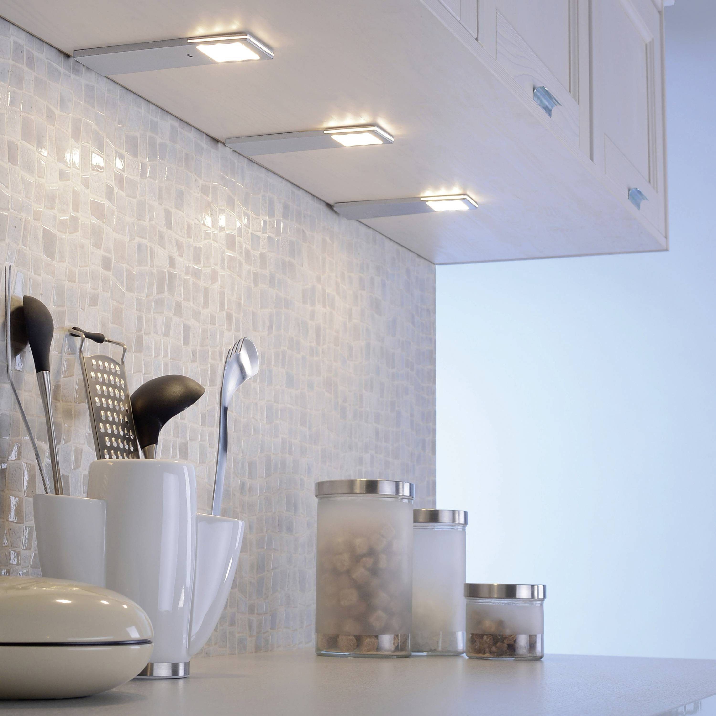 Eine Küche mit heller Arbeitsplatte, Mosaikfliesen an der Wand, Topf, Utensilienhalter und beleuchteten Hängeschränken.