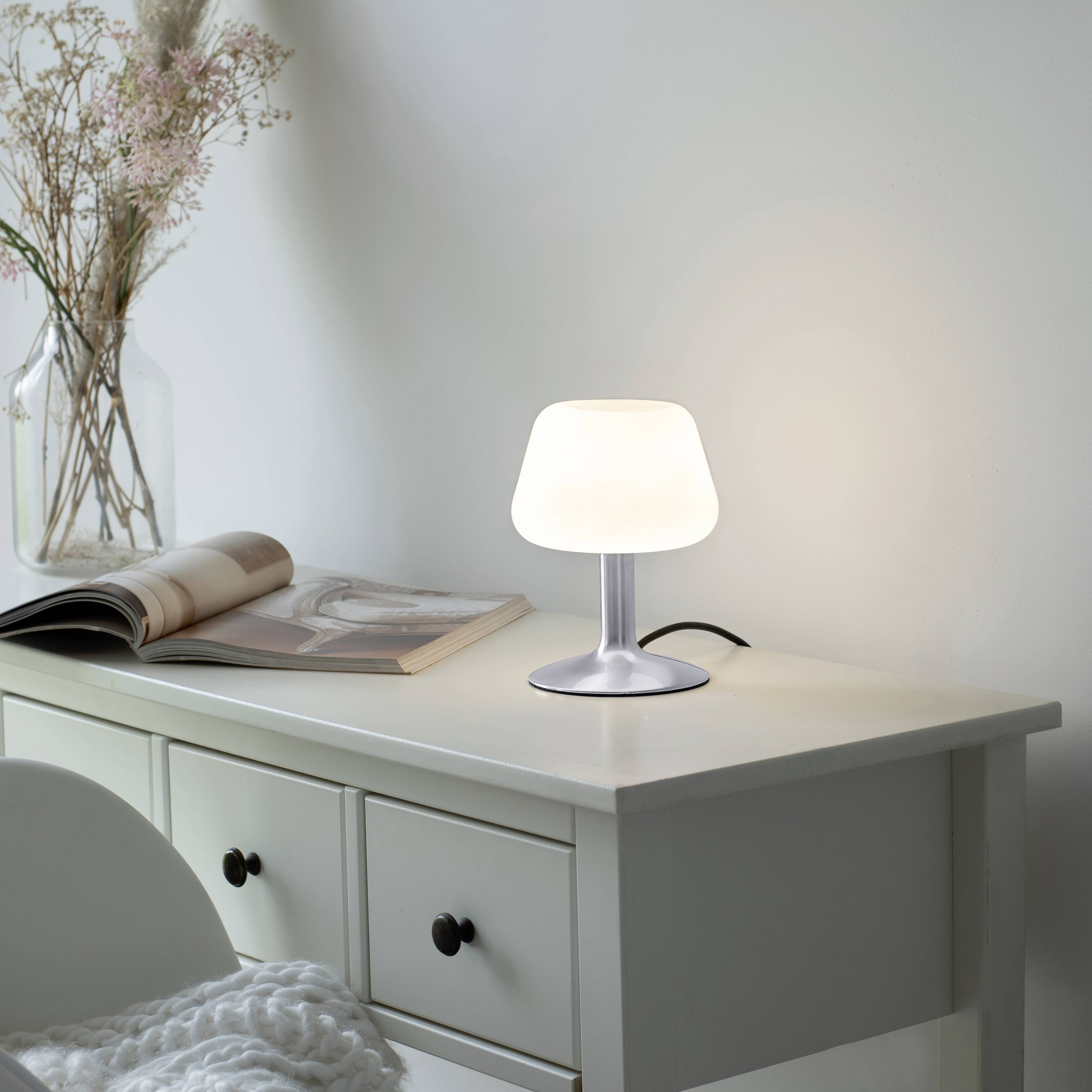 Weiße Tischlampe beleuchtet eine Kommode neben offenen Buch und Blumenvase, erzeugt eine gemütliche Atmosphäre im Raum.
