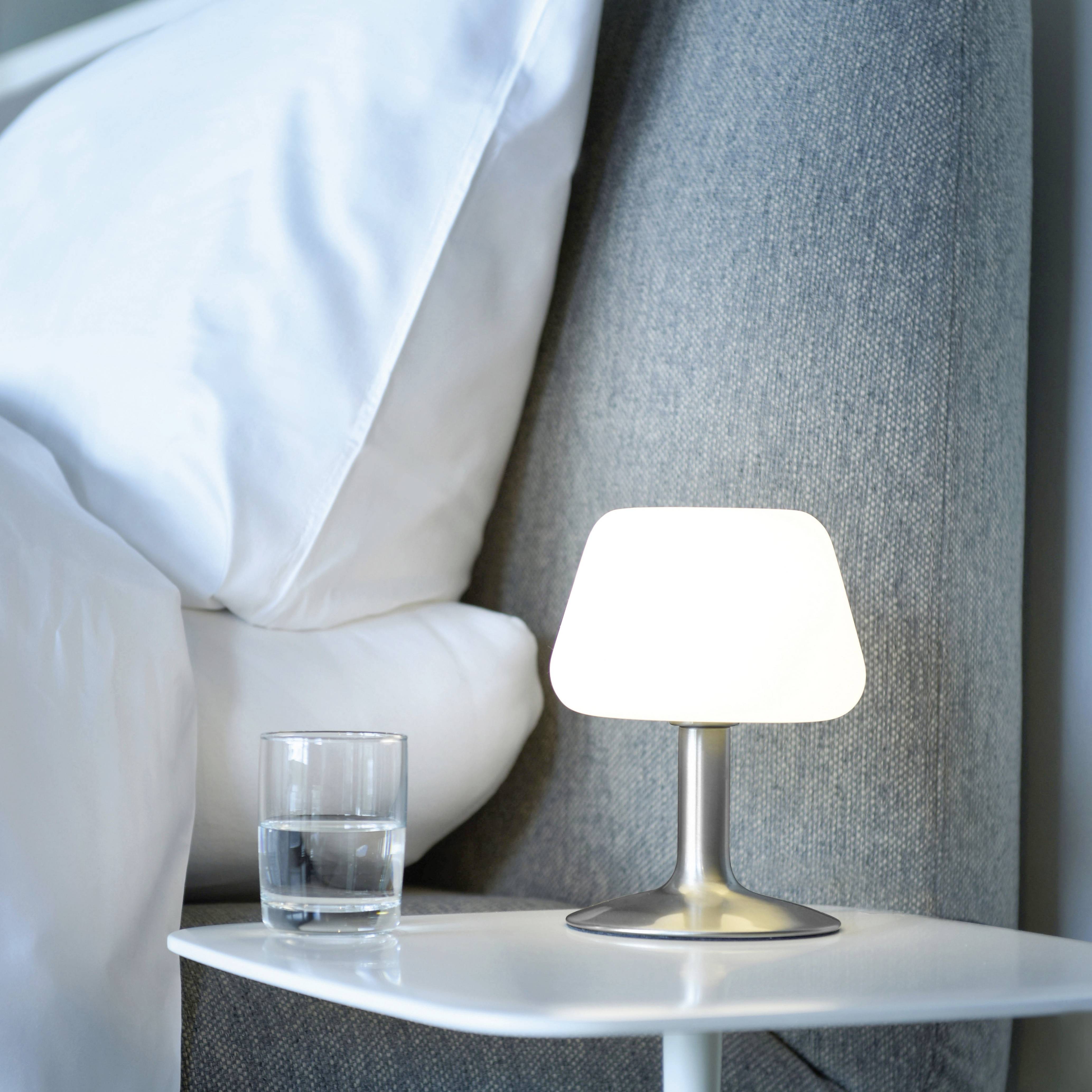 Ein Nachttisch mit einer brennenden Lampe und einem Glas Wasser steht neben einem Bett mit weißen Kopfkissen.
