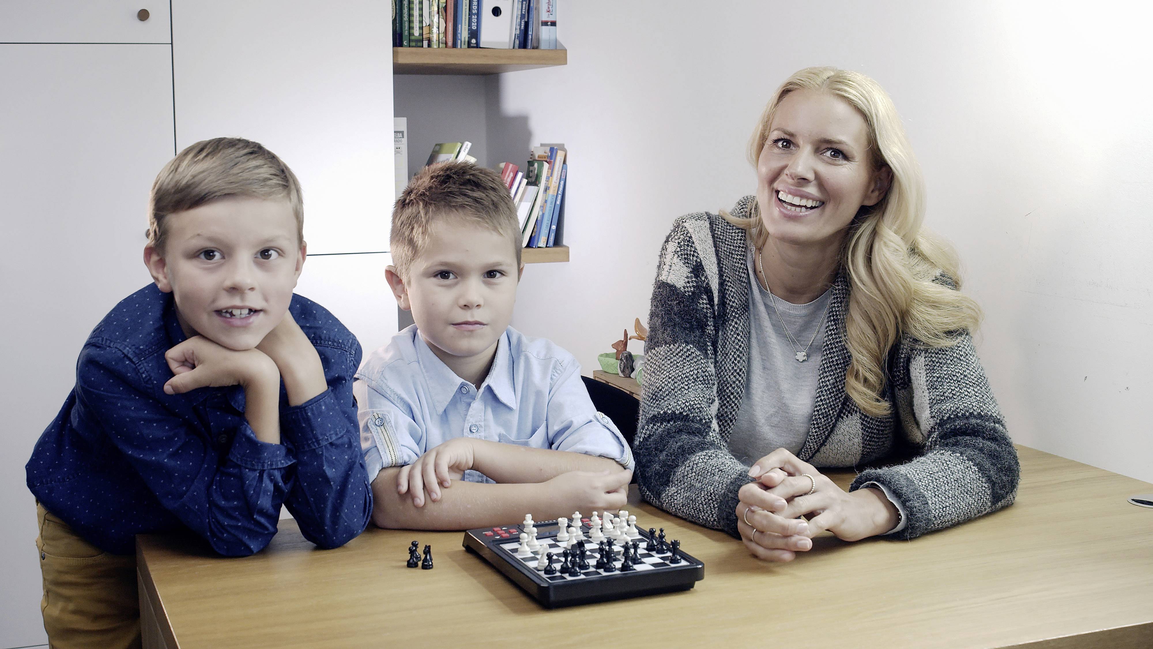 Eine Frau und zwei Jungen sitzen an einem Holztisch und spielen Schach. Die Frau lächelt, während die Jungen konzentriert schauen.