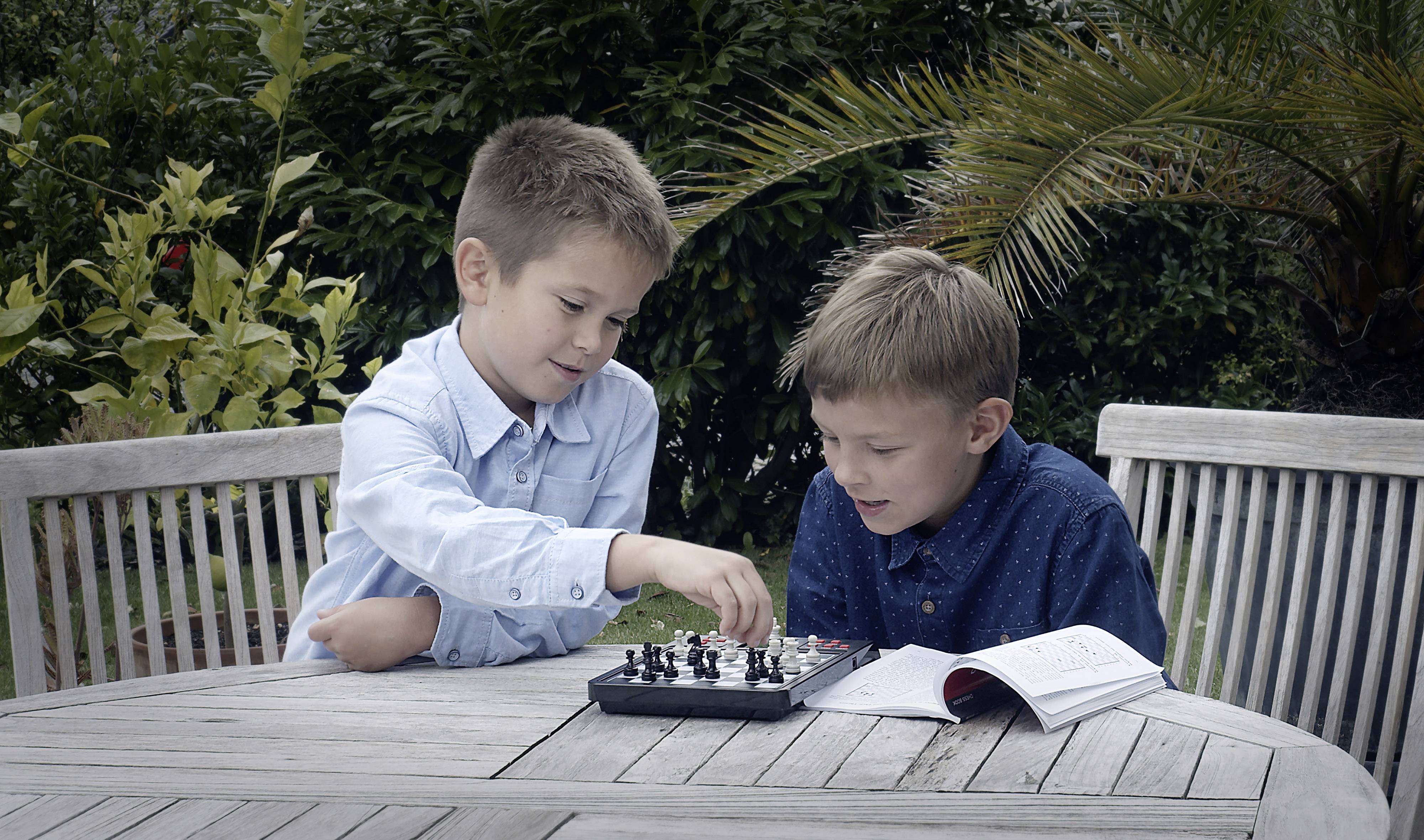 Zwei Jungen spielen draußen ein Schachspiel auf einem Tisch, umgeben von Pflanzen. Einer der Jungen macht einen Zug, während der andere zusieht.