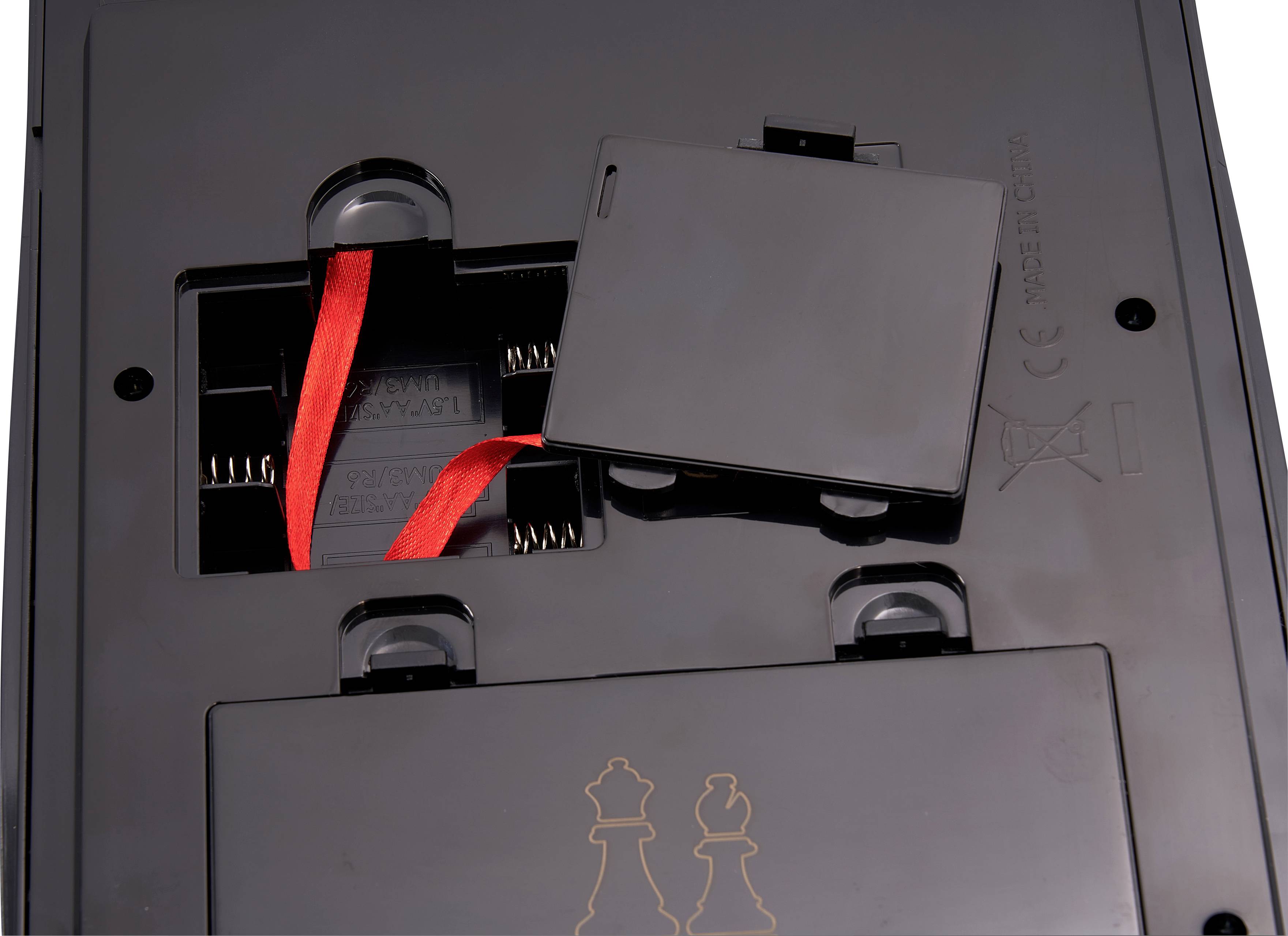 'Unterseite eines elektronischen Geräts mit offenem Batteriefach. Ein rotes Band ragt heraus. Gehäuse ist schwarz und glänzend.'