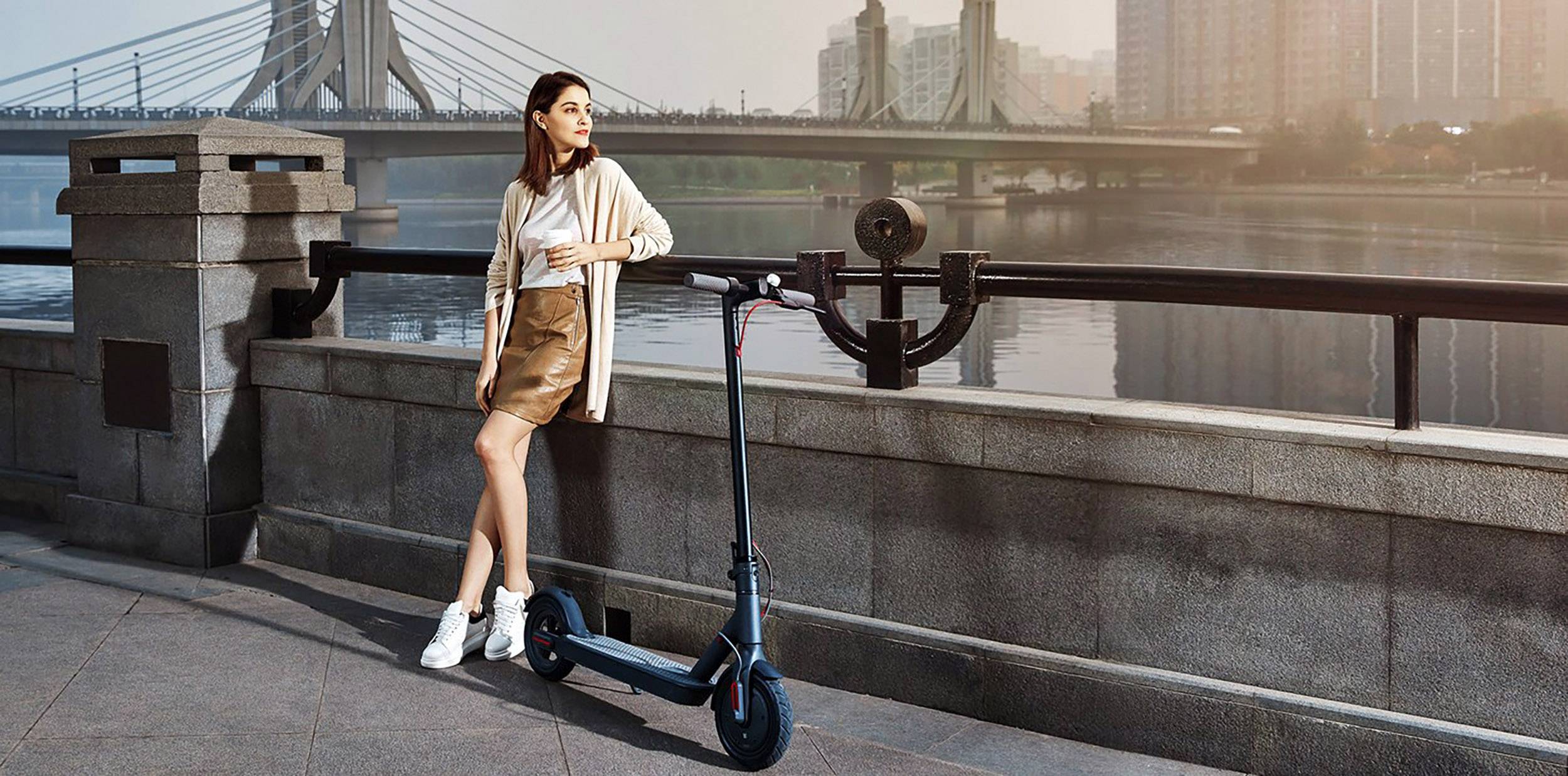 Frau lehnt an einem elektrischen Scooter und blickt über einen Fluss mit einer Brücke im Hintergrund. Städtische Umgebung bei Sonnenaufgang.