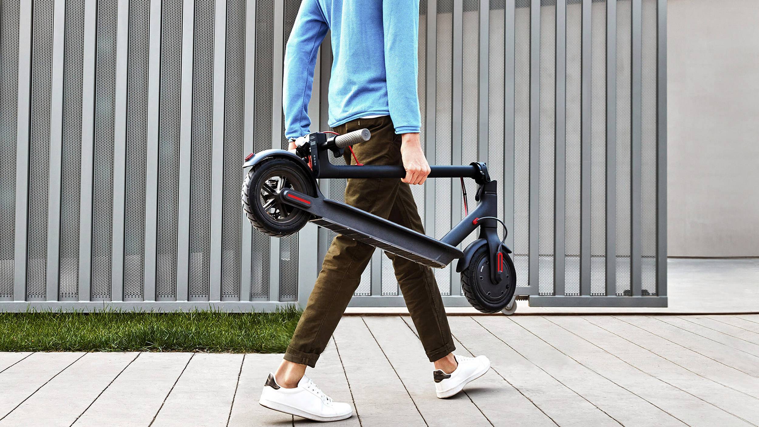 Person trägt einen zusammengeklappten E-Scooter entlang eines Gehwegs vor einer modernen, horizontal gestreiften Wand.