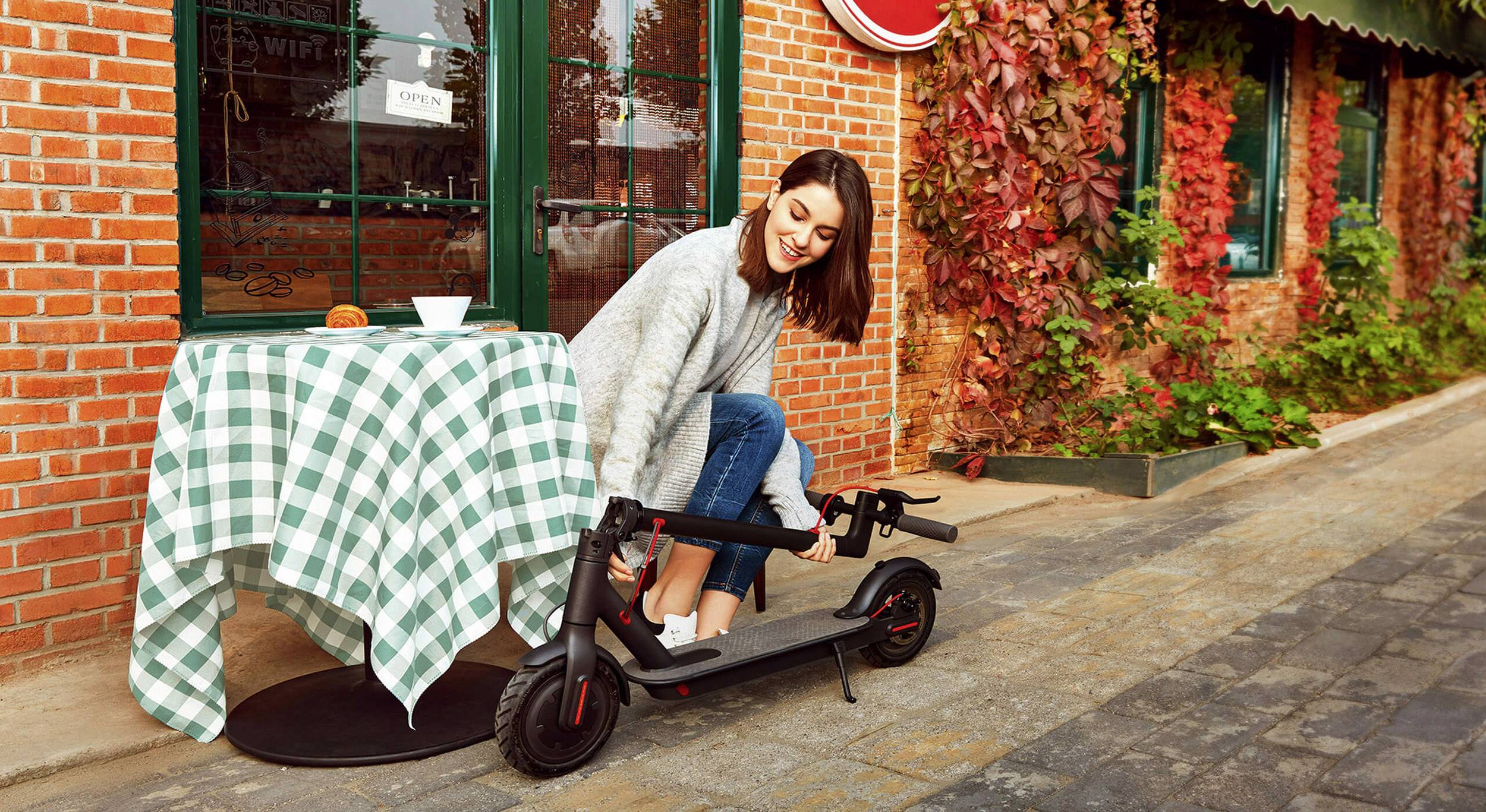 Eine Frau im Freien beugt sich, um einen E-Scooter neben einem Tisch mit kariertem Tischtuch vor einem Gebäude zu heben.