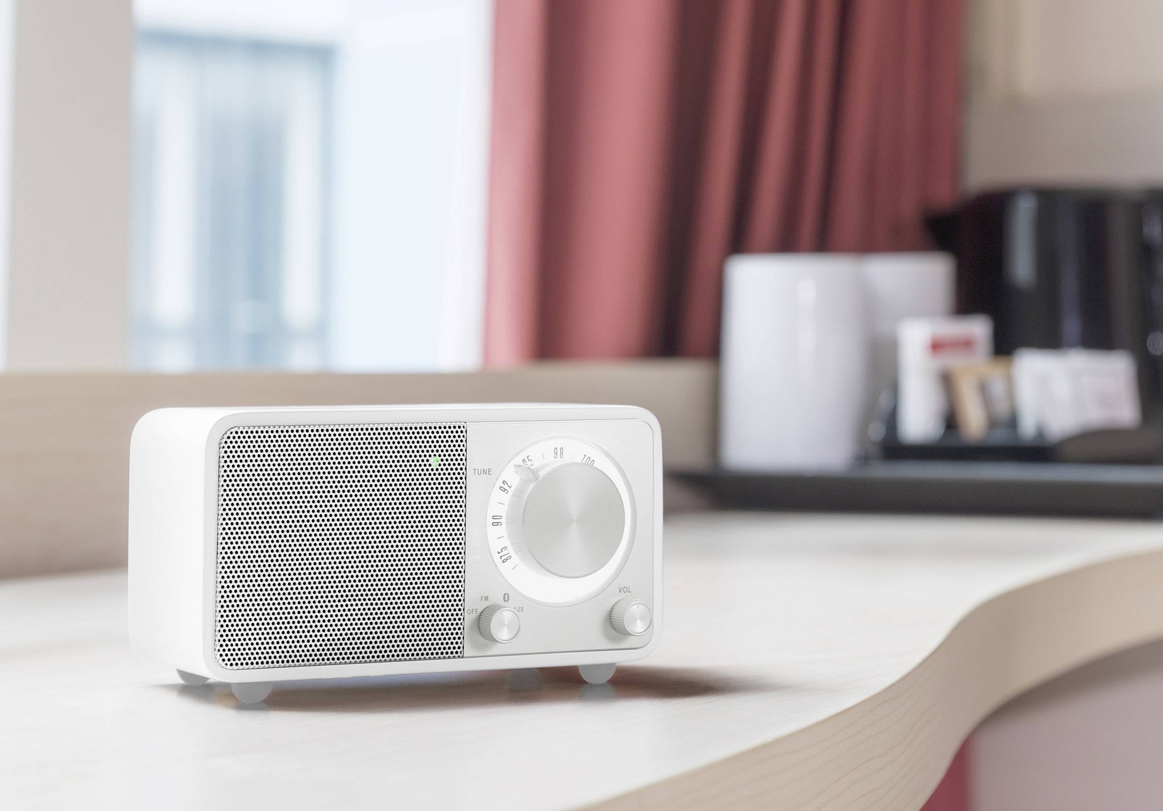 Weißes Retro-Radio mit Drehknöpfen steht auf einem Tisch vor einem Fenster. Im Hintergrund sind rote Vorhänge und eine Kaffeemaschine.