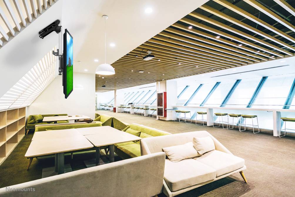 Ein moderner, gut beleuchteter Lounge-Bereich mit grünen Sofas, Tischen und einem an der Wand montierten Bildschirm. Holzbalken-Decke.