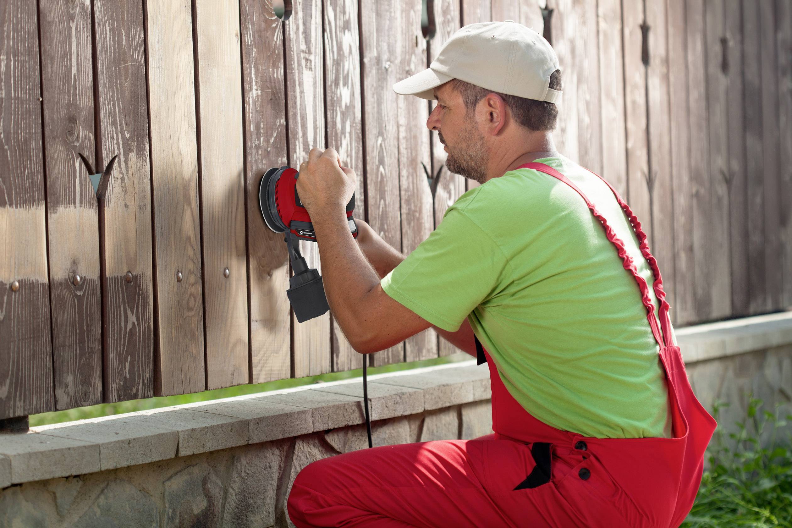 Ein Mann in roten Arbeitskleidern schleift ein Holzfurnier mit einer elektrischen Poliermaschine im Freien.