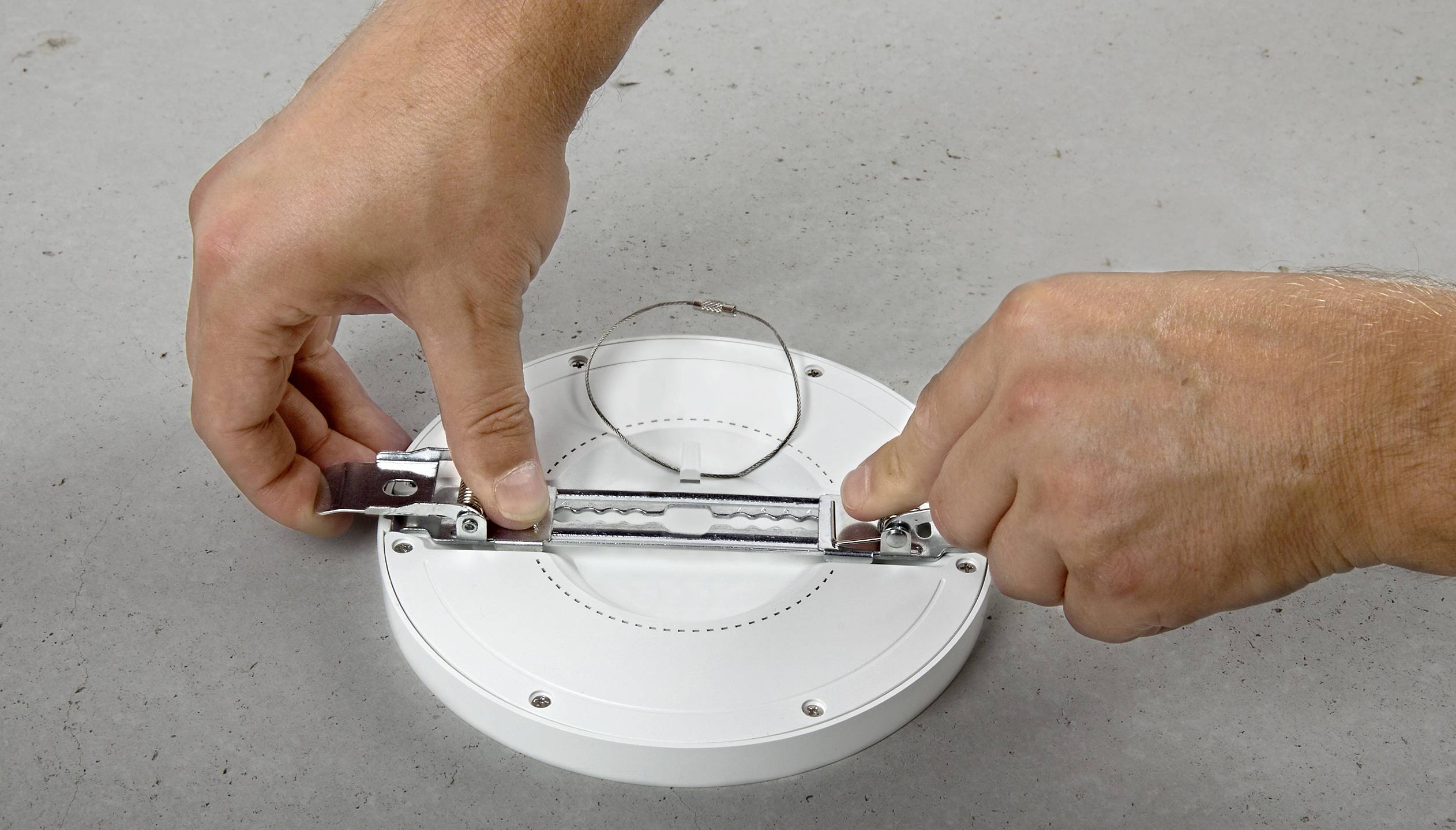Zwei Hände montieren eine runde LED-Lampe, indem sie die Halterung anbringen. Die Lampe liegt auf einem grauen Untergrund.