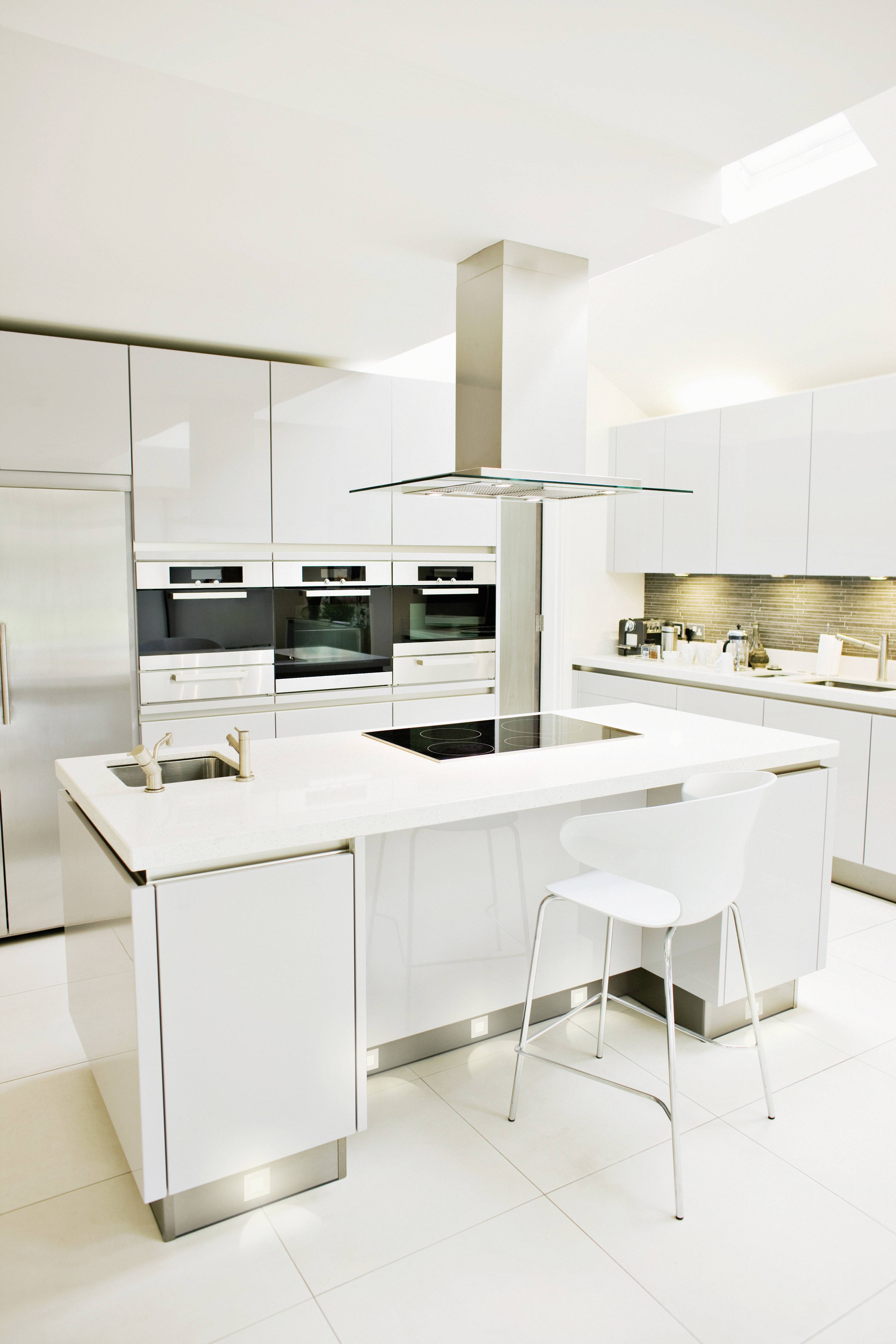 Moderne weiße Küche mit Insel, Herd, Spüle und Stuhl. Edelstahlgeräte und helle Beleuchtung betonen das minimalistische Design.