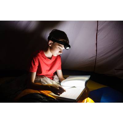 Ein Junge liest nachts unter einer Decke mit Stirnlampe ein Buch, fokussiert und interessiert.