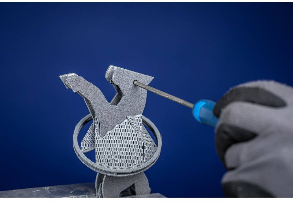 Eine Person in Handschuhen bearbeitet ein 3D-gedrucktes Objekt mit einem Werkzeug vor blauem Hintergrund.