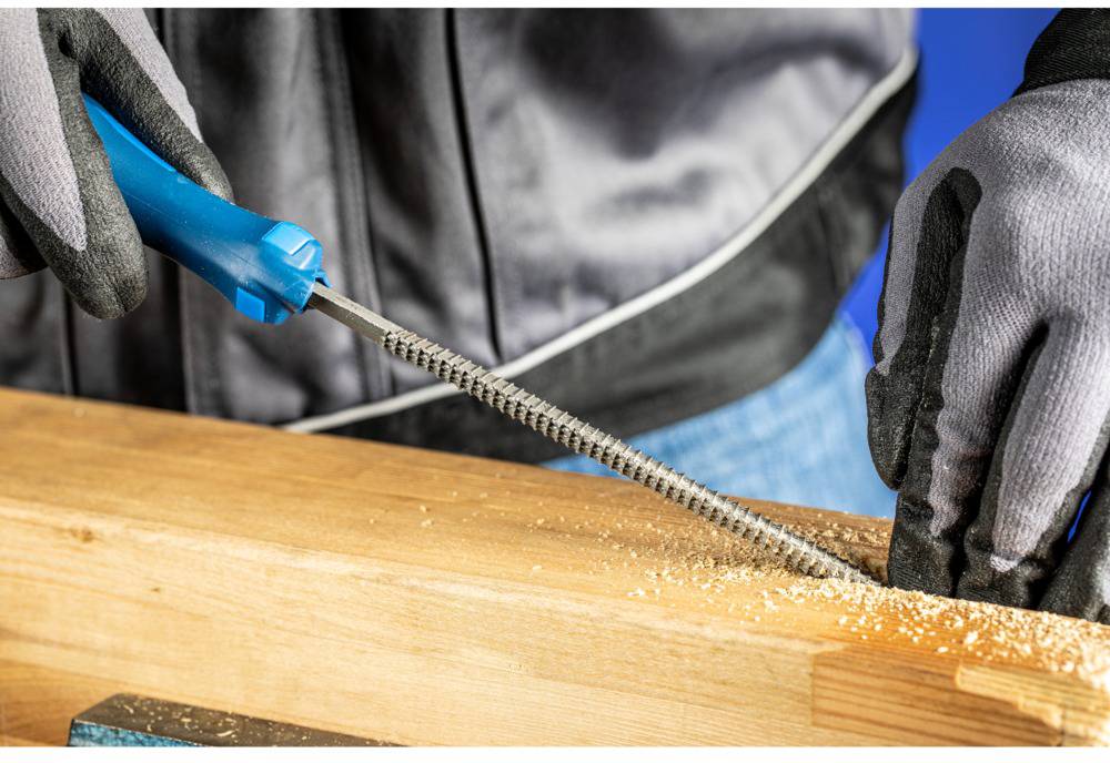 Eine Person feilt ein Holzstück mit einer Feile. Sie trägt Handschuhe, und Holzspäne sind auf der Oberfläche sichtbar.