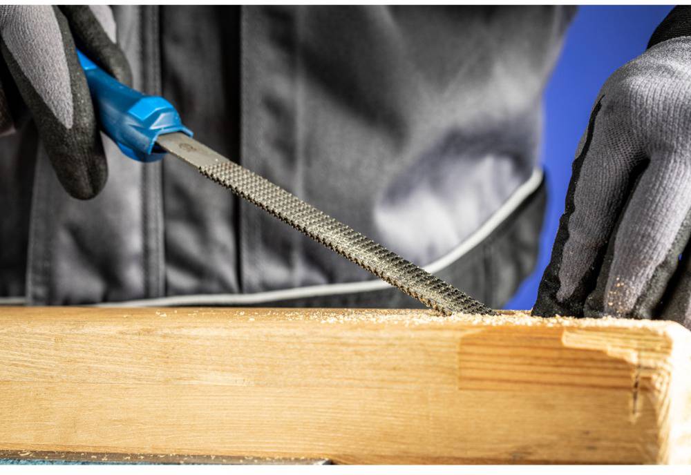 Eine Person feilt mit Handschuhen eine Holzleiste; Späne fallen. Fokus auf Feilvorgang, betont Präzision.