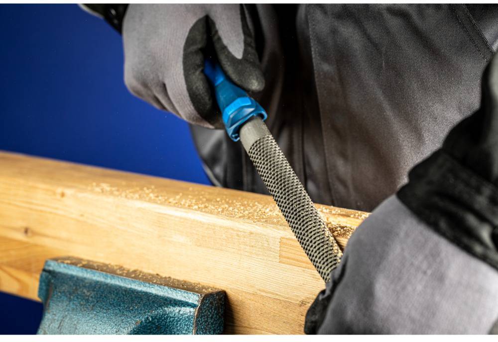Eine Person feilt Holz auf einer Werkbank. Sie trägt graue Handschuhe und eine graue Jacke. Sägespäne sind sichtbar.