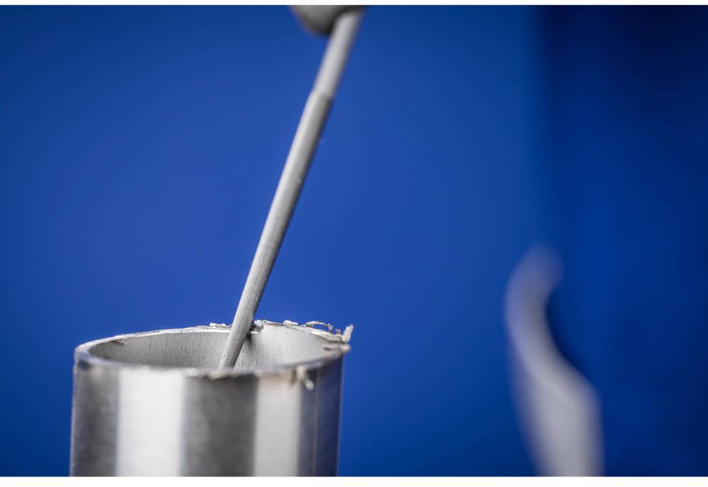 Ein Metallstab taucht in eine präzise geschnittene metallische Hülse ein vor einem blauen Hintergrund.