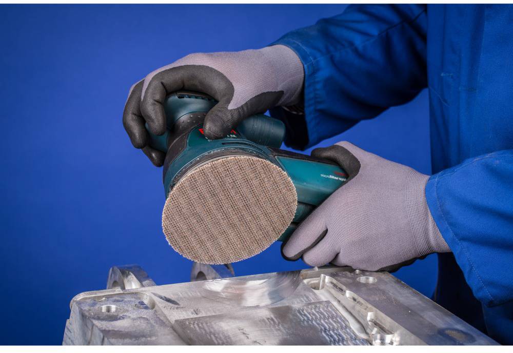 Eine Person schleift eine Metalloberfläche mit einem elektrischen Winkelschleifer in einer Werkstattumgebung. Sie trägt Sicherheitshandschuhe.