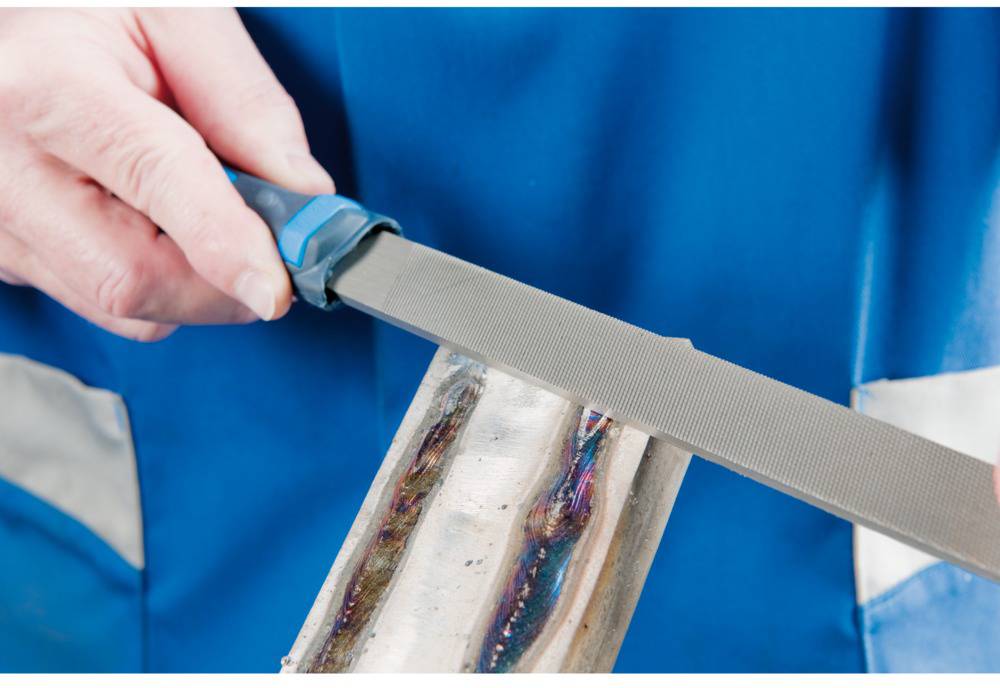 Eine Person feilt Metall mit einer Metallfeile, gekleidet in blauer Arbeitskleidung. Die Feile bearbeitet eine Kante.