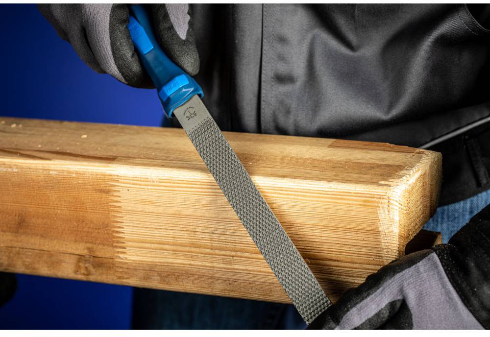 Eine Person feilt ein Holzstück mit einer blauen Feile, trägt dabei Handschuhe und eine schwarze Jacke.