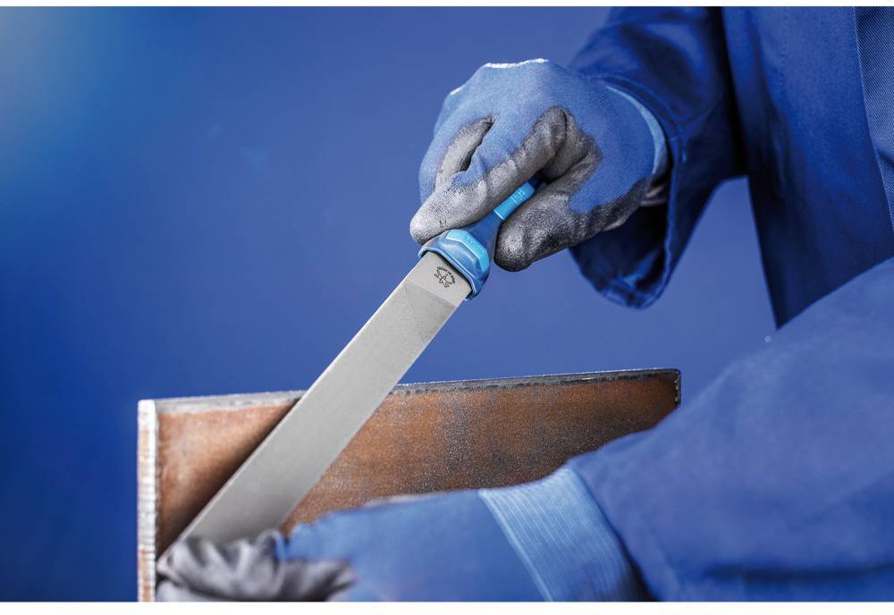 'Nahaufnahme einer Person, die mit einer Feile eine Metallkante bearbeitet, trägt blaue Arbeitskleidung und Schutzhandschuhe.'