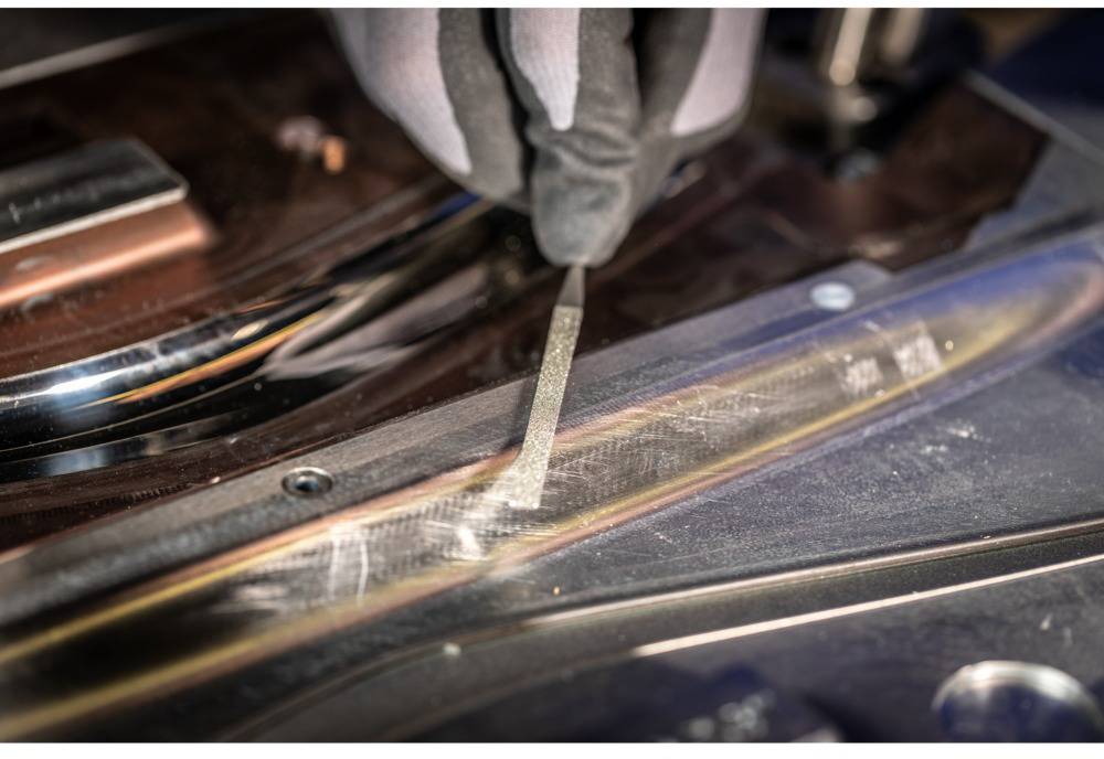 'Eine Hand in einem Handschuh bearbeitet eine Metalloberfläche mit einem Werkzeug, vermutlich bei einem industriellen Fertigungsprozess.'