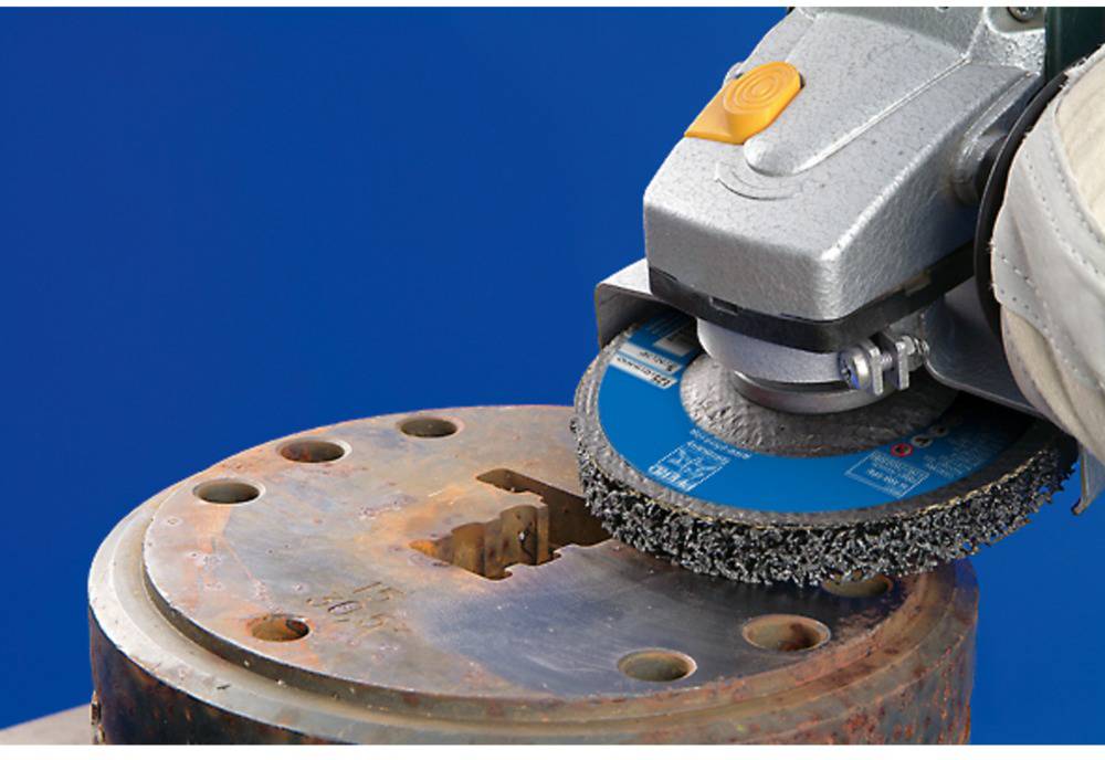 Eine Schleifmaschine bearbeitet die Oberfläche eines rostigen Metallteils vor blauem Hintergrund.