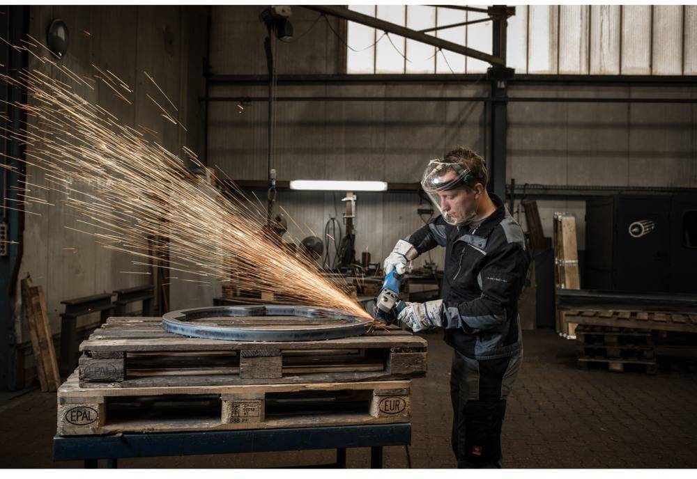 Eine Person in Schutzkleidung schleift mit einem Winkelschleifer Metall, Funken sprühen. Industriewerkstatt im Hintergrund.