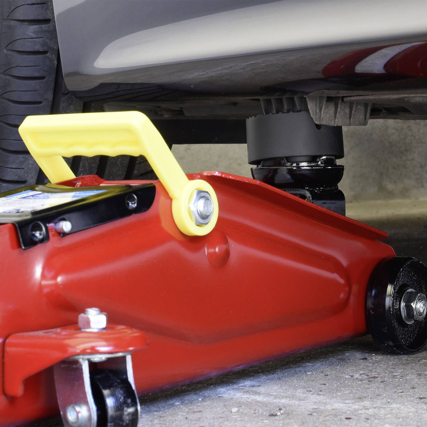 Ein Autoheber hebt ein Auto in einer Garage. Der Wagenheber ist rot mit einem gelben Griff und befindet sich unter einem silbernen Auto.