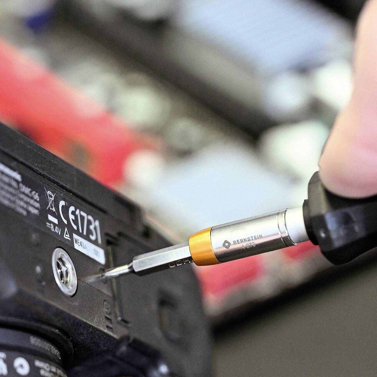 Eine Person benutzt einen Schraubendreher, um eine Schraube an einem elektronischen Gerät anzuziehen, vor einem unscharfen Leiterplattenhintergrund.