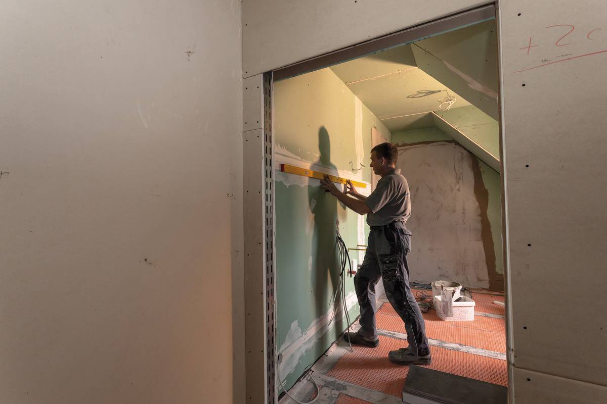 Ein Bauarbeiter verwendet eine Wasserwaage, um eine Wand in einem unfertigen Raum zu nivellieren. Die Umgebung ist noch im Rohbau.