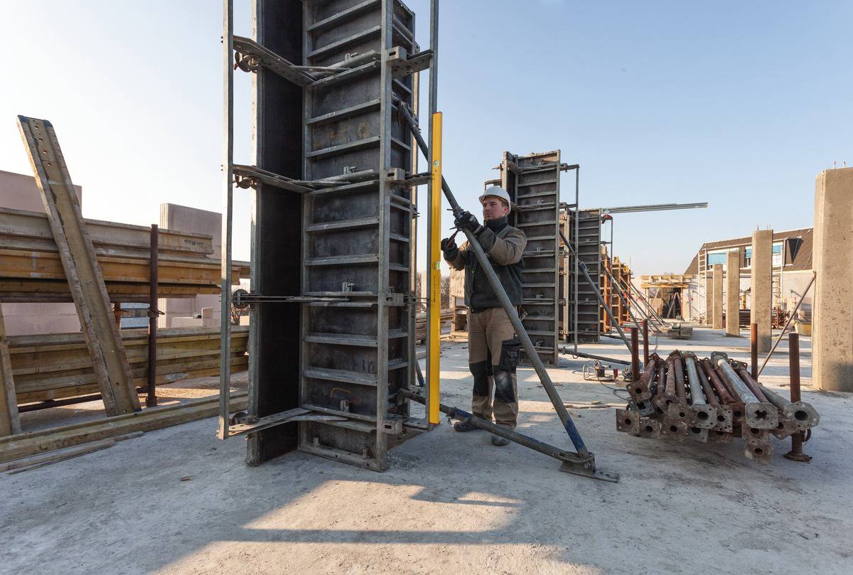 Ein Bauarbeiter befestigt eine Schalungsstütze an einer Betonform auf einer Baustelle. Um ihn herum stehen verschiedene Bauformen.