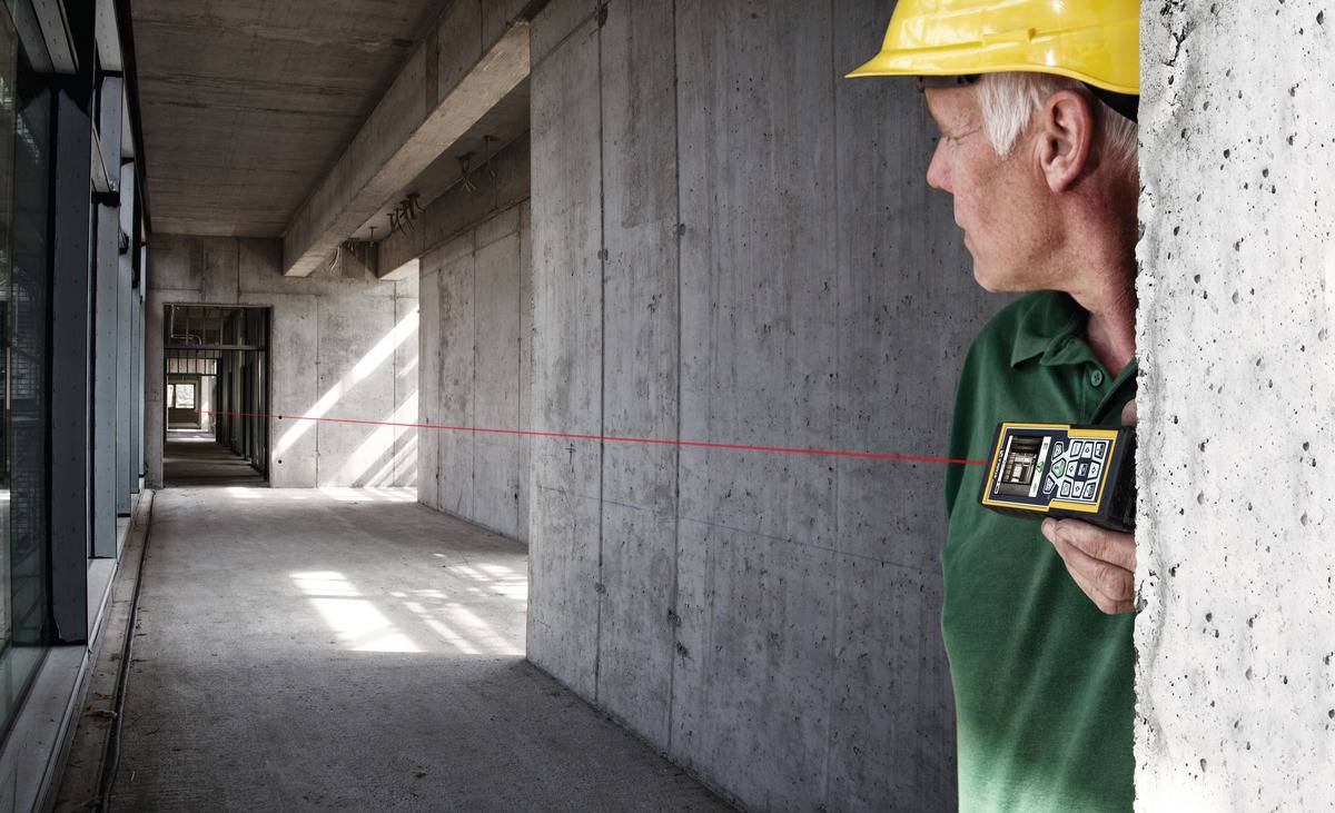 Ein Bauarbeiter in einem gelben Schutzhelm misst eine lange Betonhalle mit einem Laser-Distanzmesser.