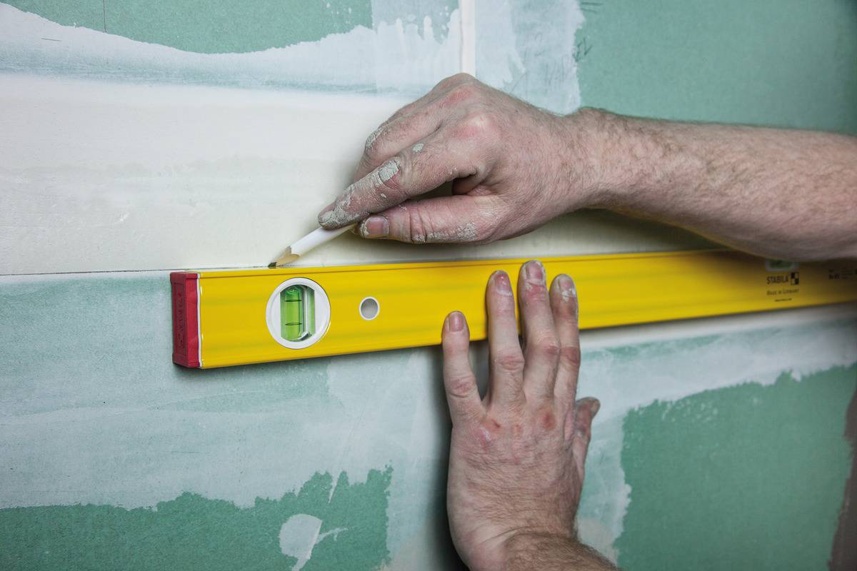 'Eine Person zeichnet mit einem Bleistift eine Linie an der Wand entlang einer gelben Wasserwaage, um korrekte Ausrichtung sicherzustellen.'